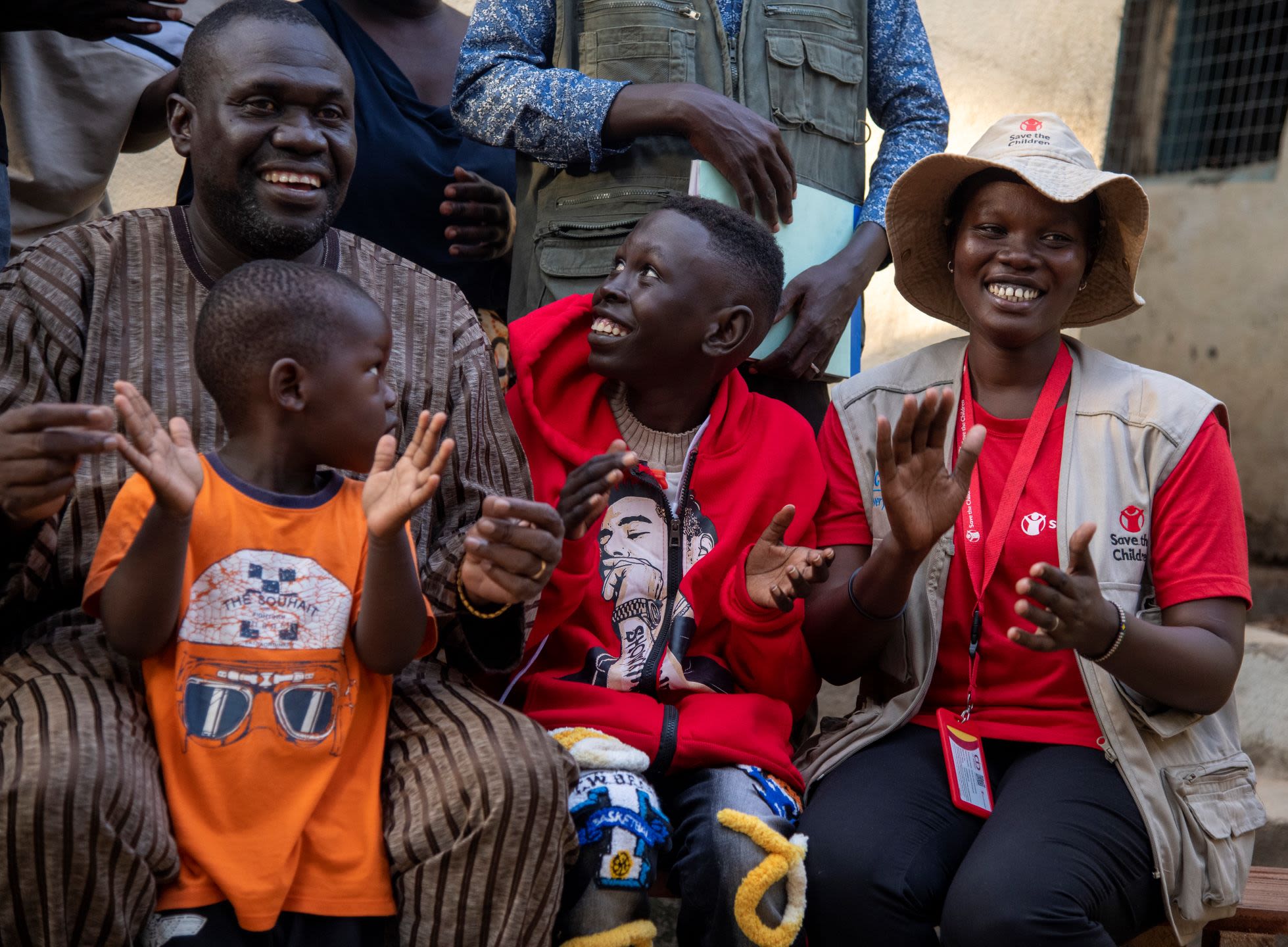 Photo: Simon*, 13, wears a red jumper and smiles and claps with family members after they were reunited in South Sudan. Credit: Cicilia Ade Hillary/ Save the Children.