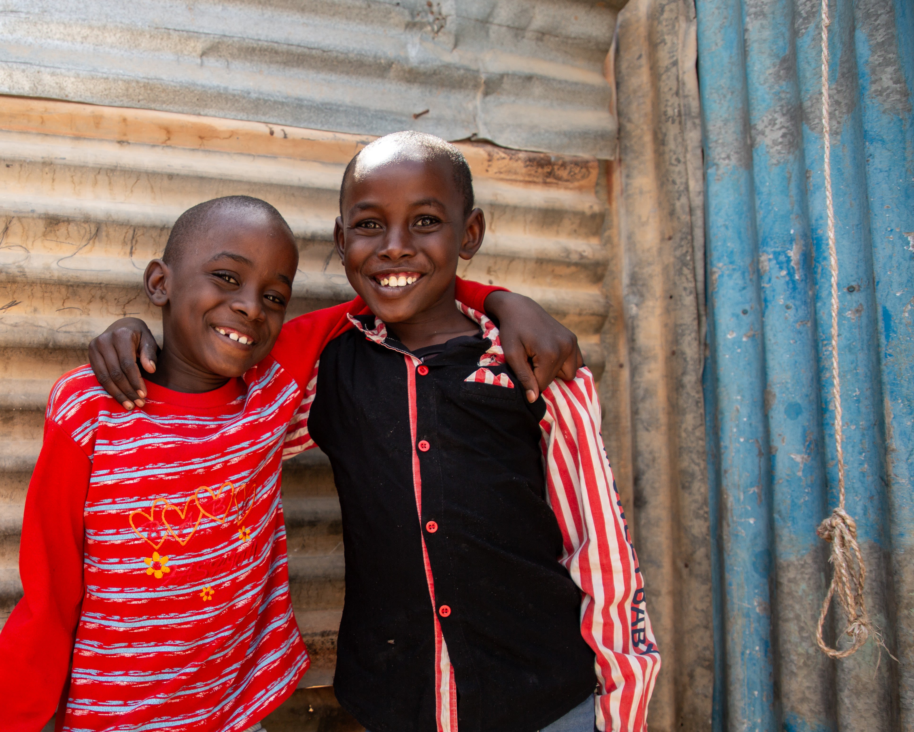 Photo: Omar, 7, wearing red, with his brother, wearing black, both smiling at the camera at the Internally Displaced Persons (IDP) camp where they live, Somalia. Credit: Mustafa Saeed / Save the Children.