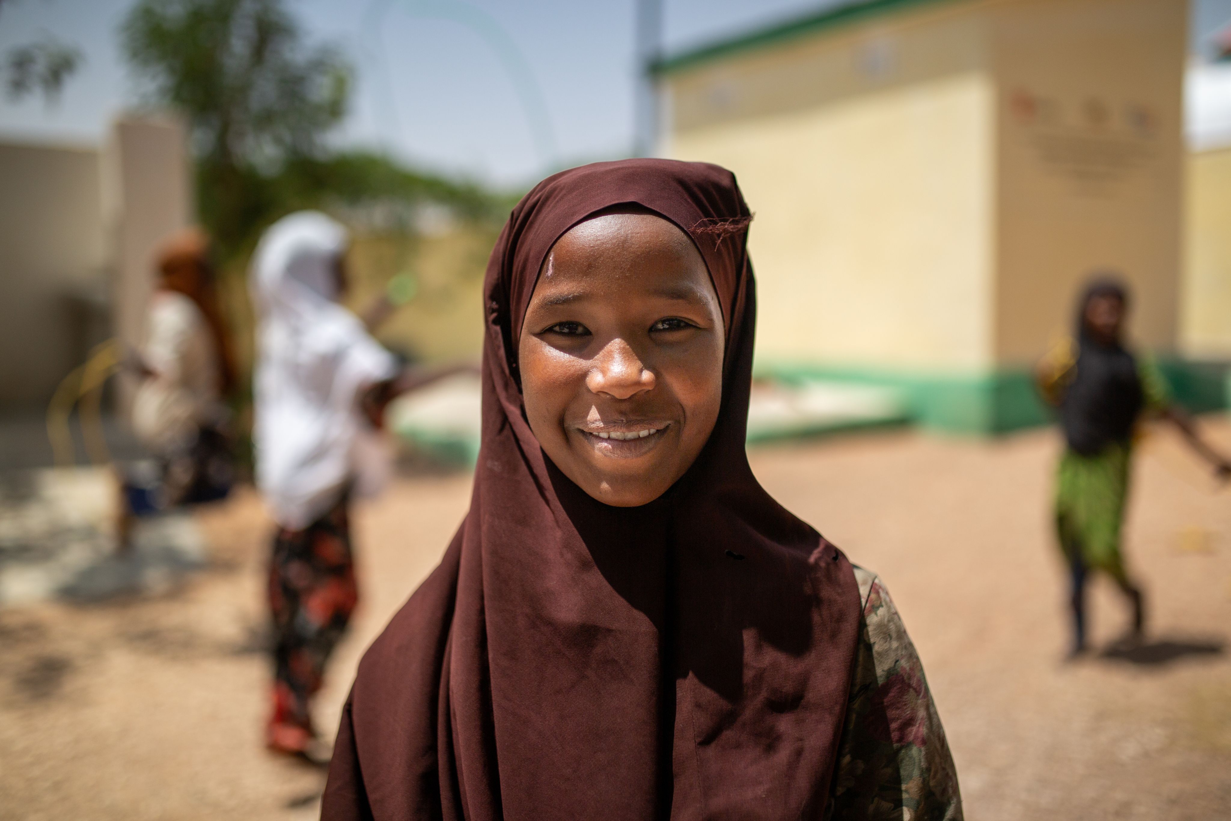 Faisa*, a girl of 12, wearing a brown hijab and smiling, playing outside at a displaced persons camp, Somalia.